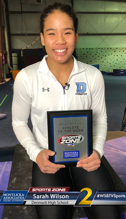 Young female gymnast holding Sports Zone Athlete of the Week award, smiling at camera in gymnasium.