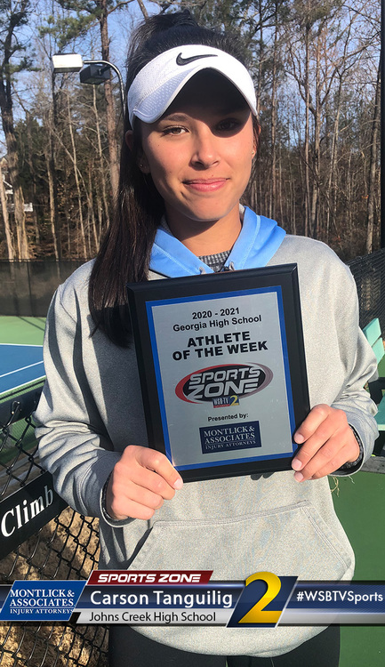 Young female student athlete of the week holding award plaque on tennis court, smiling at camera.