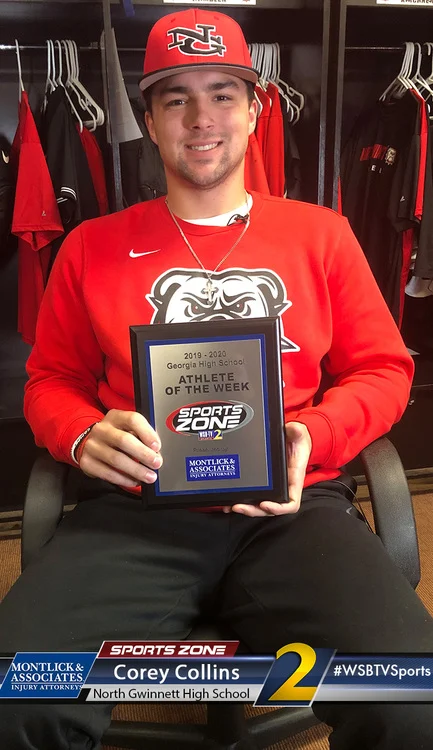 Young male athlete smiling and holding "Athlete of the Week" sports award plaque while sitting in locker room.