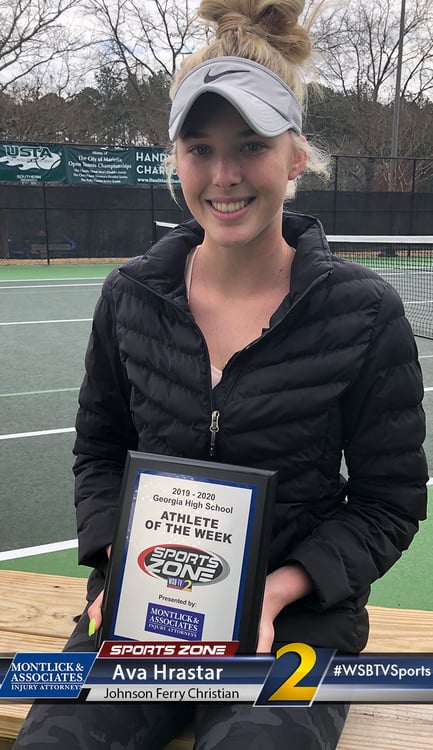 Smiling female athlete of the week holding award plaque on tennis court, wearing black Nike jacket