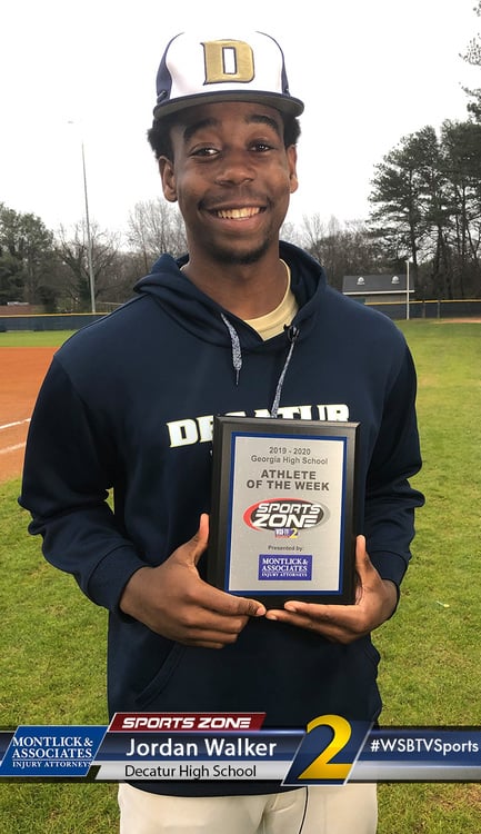 Smiling high school athlete holding "Athlete of the Week" plaque on baseball field with trees in background.