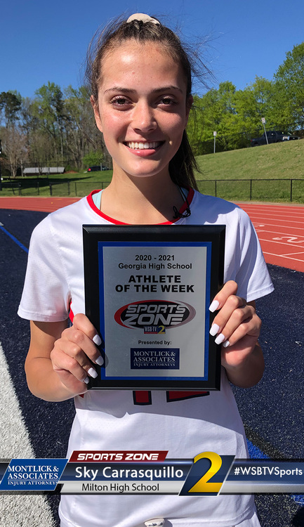 Smiling high school athlete of the week holding award plaque on running track at school sports complex.