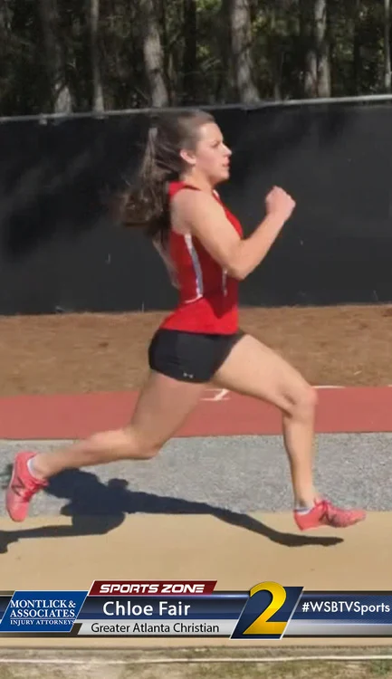 Female athlete lunging in red tank top and shorts at outdoor track, raising fist in running motion