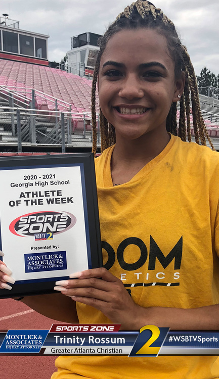 Young female athlete smiling while holding "Athlete of the Week" award at high school stadium track.