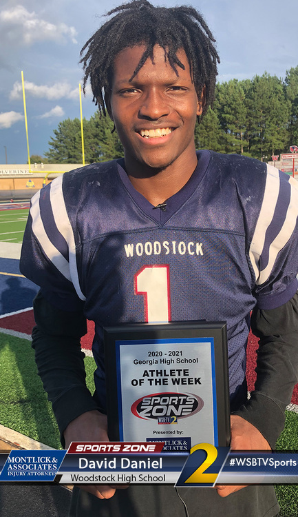 Smiling high school football player holding Athlete of the Week award on football field in jersey.