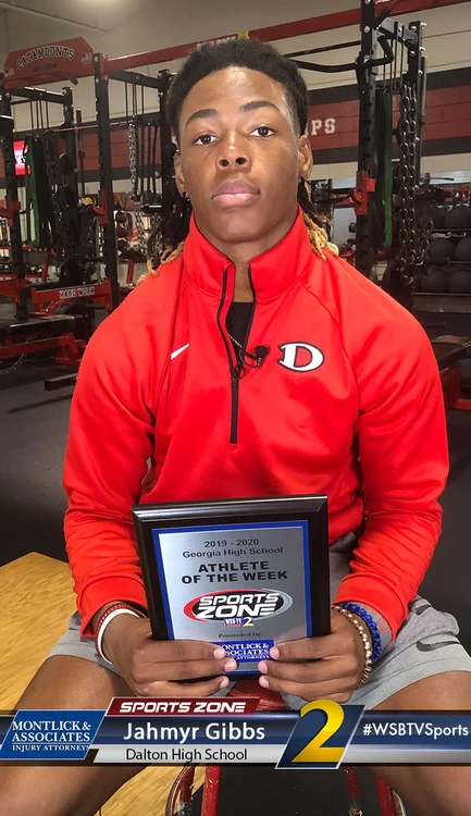Dalton High School athlete of the week Jahmyr Gibbs holds Sports Zone award in red athletic gear at gym.