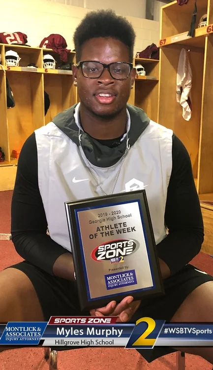 Student athlete of the week smiles while holding award plaque in locker room, wearing glasses and hoodie.