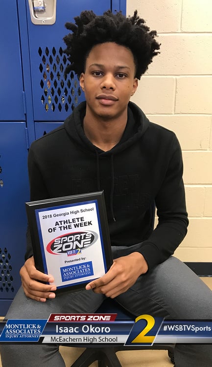 Young African American male athlete holding Sports Zone Athlete of the Week award plaque at high school locker. The image shows a smiling young man with an afro hairstyle proudly displaying his athletic achievement award in front of blue lockers, likely at his school.