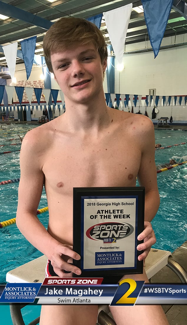 Shirtless male high school athlete holding "Athlete of the Week" plaque near swimming pool lanes and banners.