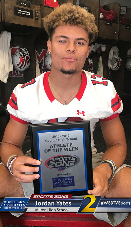 High school football player holding SportsZone Athlete of the Week award in locker room, smiling at camera.