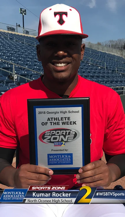 Smiling athlete in red uniform holds Athlete of the Week plaque at sports stadium bleachers.