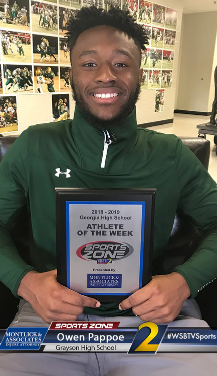 Smiling athlete of the week award winner in green jacket holding plaque, wall of sports photos behind him