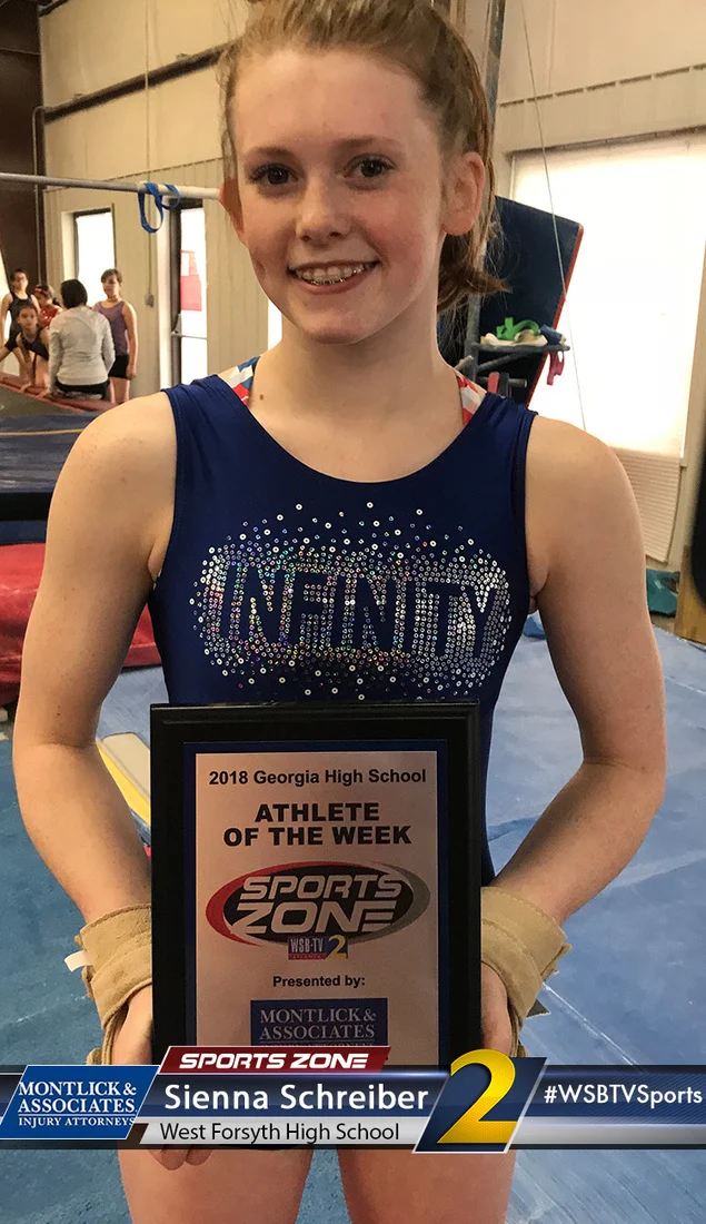 Young female gymnast posing with "Athlete of the Week" award plaque smiling at a gymnastics gym.