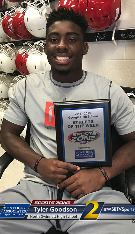 North Gwinnett High School athlete of the week Tyler Goodson smiles while holding his award plaque in front of a display of football helmets.