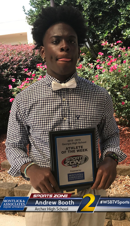 Athlete of the Week award winner holding plaque, standing in front of blooming flowers, smiling proudly