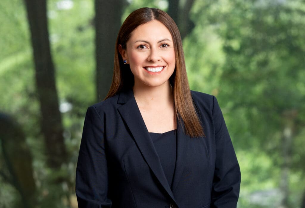 Smiling female attorney in navy suit with brown hair, outdoors with trees in background