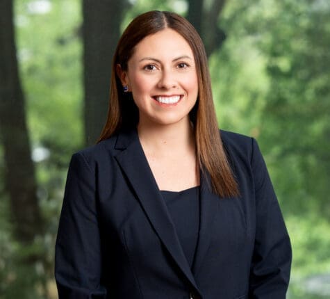Smiling female attorney in navy suit with brown hair, outdoors with trees in background