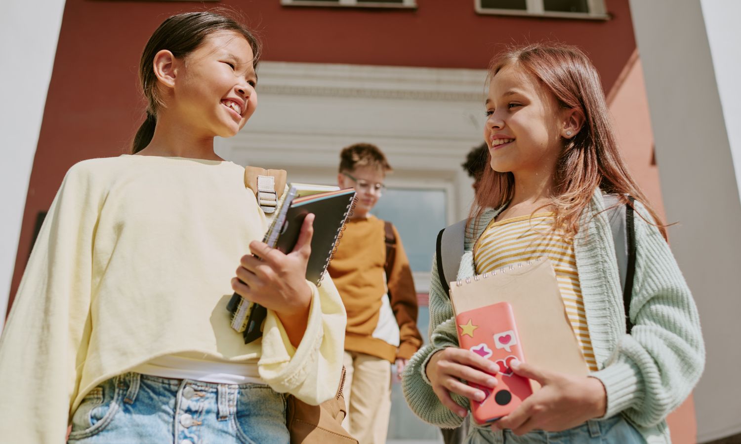 two preteen girls outside of a school, holding notebooks and mobile phones